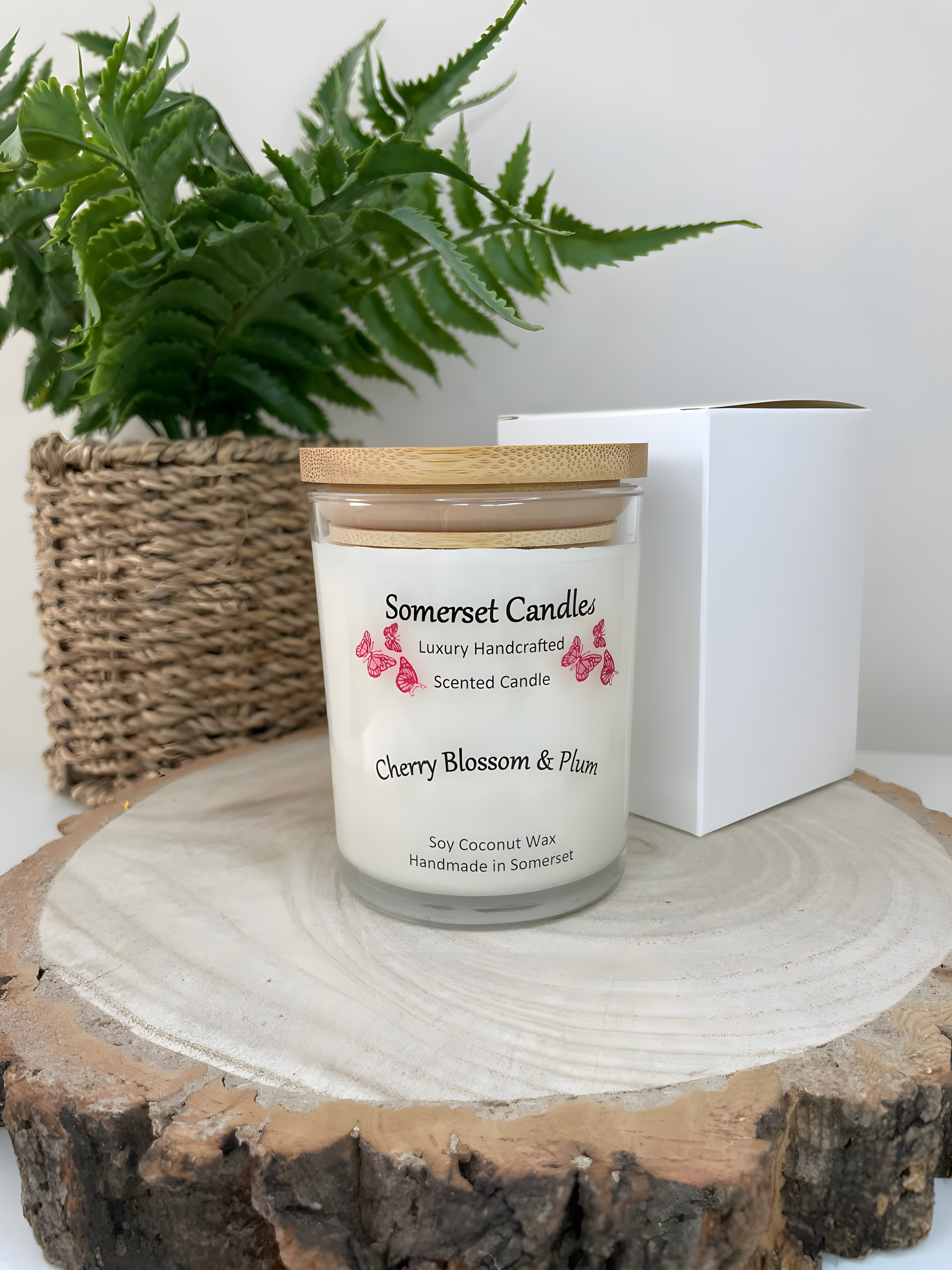 Cherry blossom & plum scented soy candle is displayed on a wooden board with a white gift box and a plant in the back ground. The candle is white in colour with bright red butterflies above the scent name with a lovely bamboo lid.