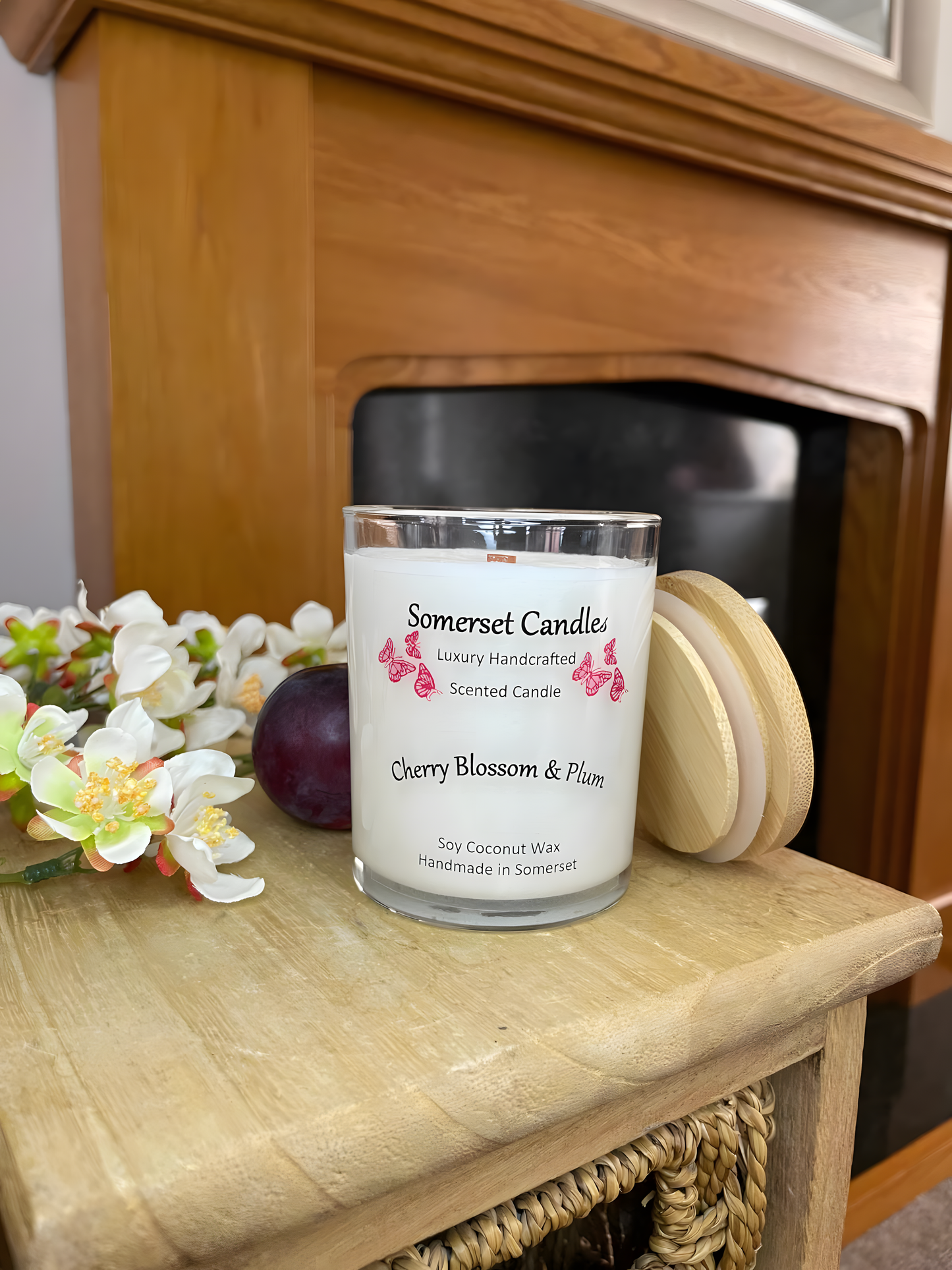 Cherry blossom & plum scented soy candle is displayed on a wooden board with a fresh plum & white cherry blossom in front of a fire place. The candle is white in colour with bright red butterflies above the scent name with a bamboo lid at the side.