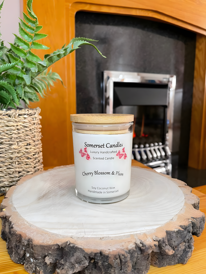 Cherry blossom & plum scented soy candle is displayed on a wooden board with a plant in front of a lit fire place. The candle is white in colour with bright red butterflies above the scent name with a lovely bamboo lid fitted.