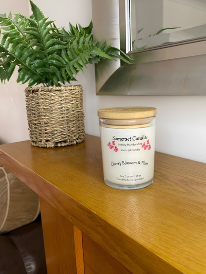 Cherry blossom & plum scented soy candle is displayed on a wooden mantel piece with a plant behind. The candle is white in colour with bright red butterflies above the scent name with a lovely bamboo lid fitted.