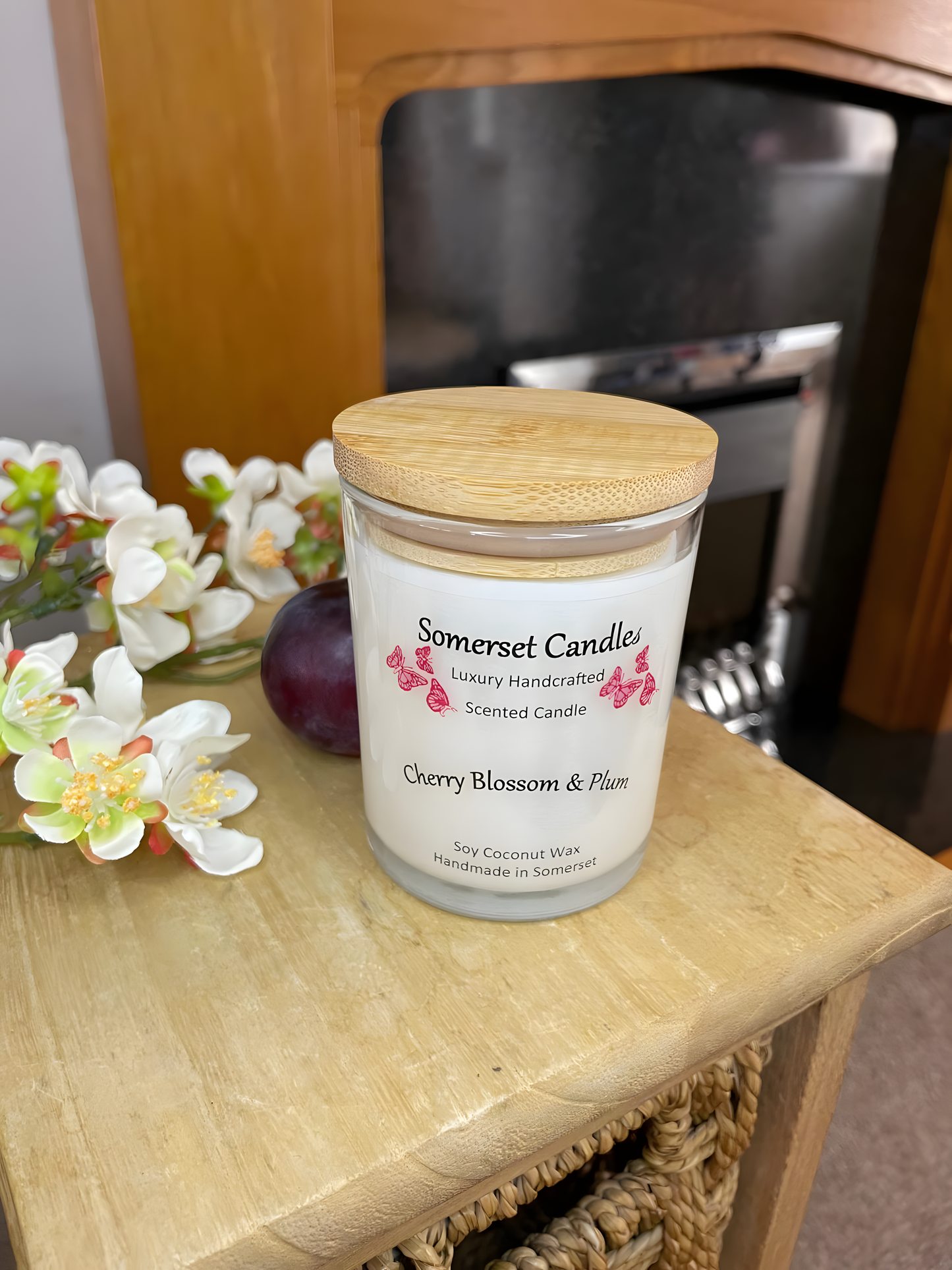 Cherry blossom & plum scented soy candle is displayed on a wooden board with a fresh plum & white cherry blossom in front of a fire place. The candle is white in colour with bright red butterflies above the scent name with a lovely bamboo lid.