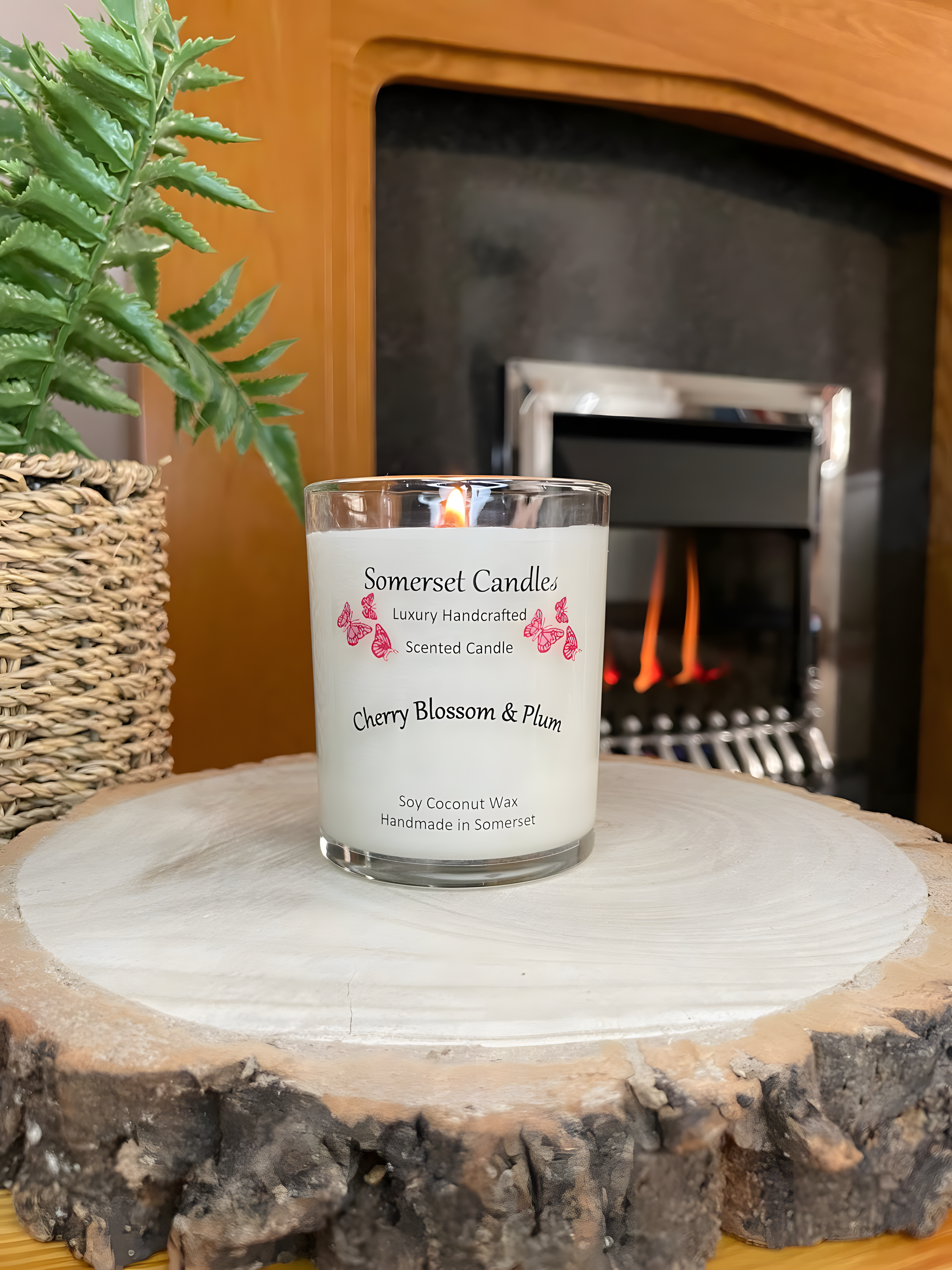 Cherry blossom & plum scented soy candle is displayed on a wooden board with a plant and in front of a lit fire. The candle is white in colour with bright red butterflies above the scent name. The candle is lit.