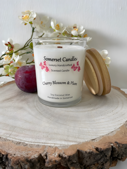 Cherry blossom & plum scented soy candle is displayed on a wooden board with a fresh plum & white cherry blossom. The candle is white in colour with bright red butterflies above the scent name with a lovely bamboo lid lent to the right of the candle.