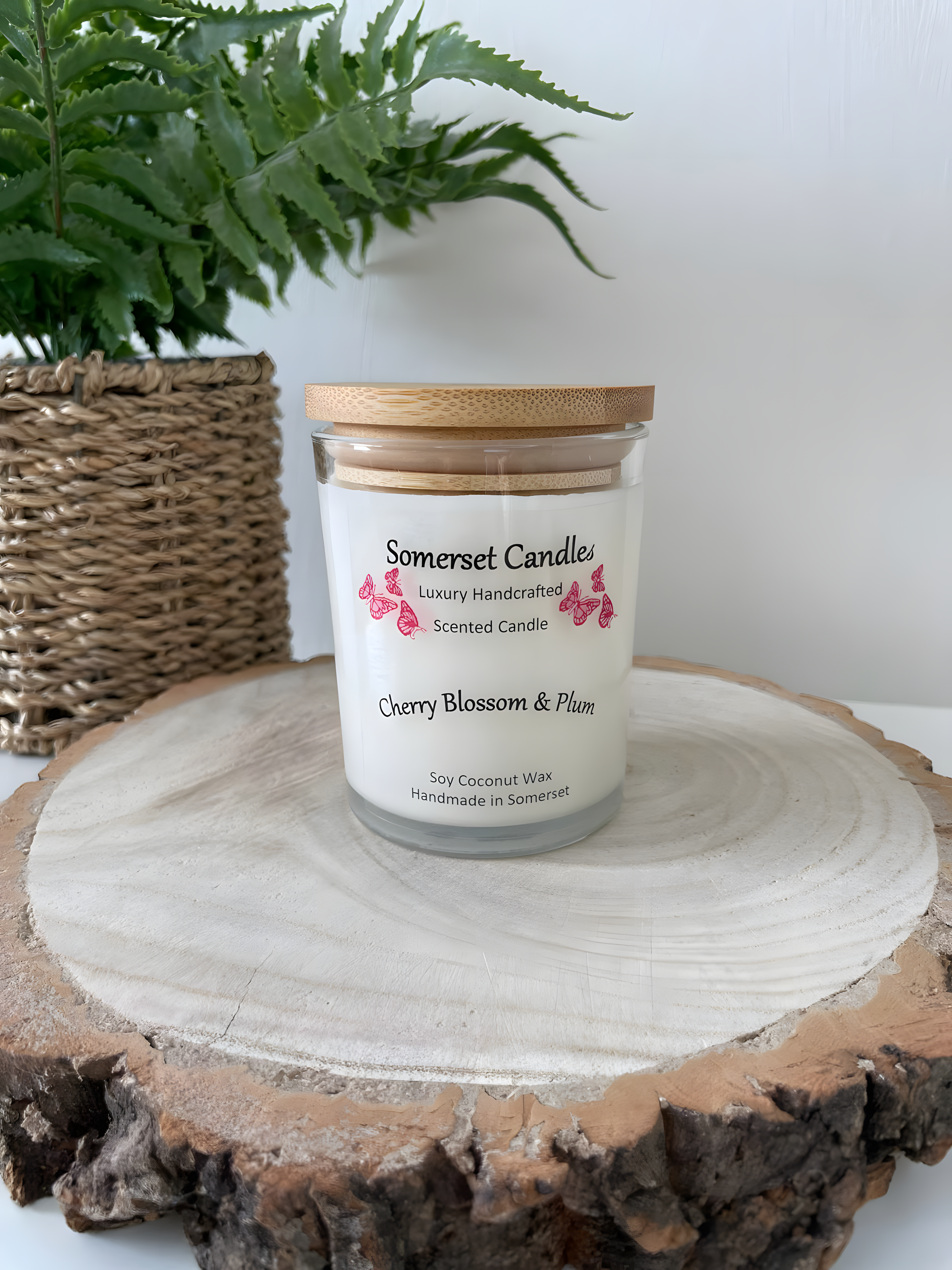 Cherry blossom & plum scented soy candle is displayed on a wooden board with plant behind. The candle is white in colour with bright red butterflies above the scent name with a lovely bamboo lid fitted.