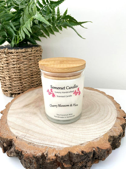 White soy candle in a 7oz clear glass jar with a natural wooden lid. The candle is made from a soy-coconut blend and scented with a delicate mix of cherry blossom and plum. Above the scent name on the label, several red butterflies are displayed, adding a romantic touch. Clean, minimalist design ideal for gifts, home decor or self-care.