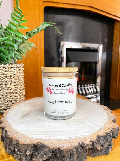 White soy candle in a 7oz clear glass jar with a natural wooden lid. The candle is made from a soy-coconut blend and scented with a delicate mix of cherry blossom and plum. Above the scent name on the label, several red butterflies are displayed, adding a romantic touch. Clean, minimalist design ideal for gifts, home decor or self-care.