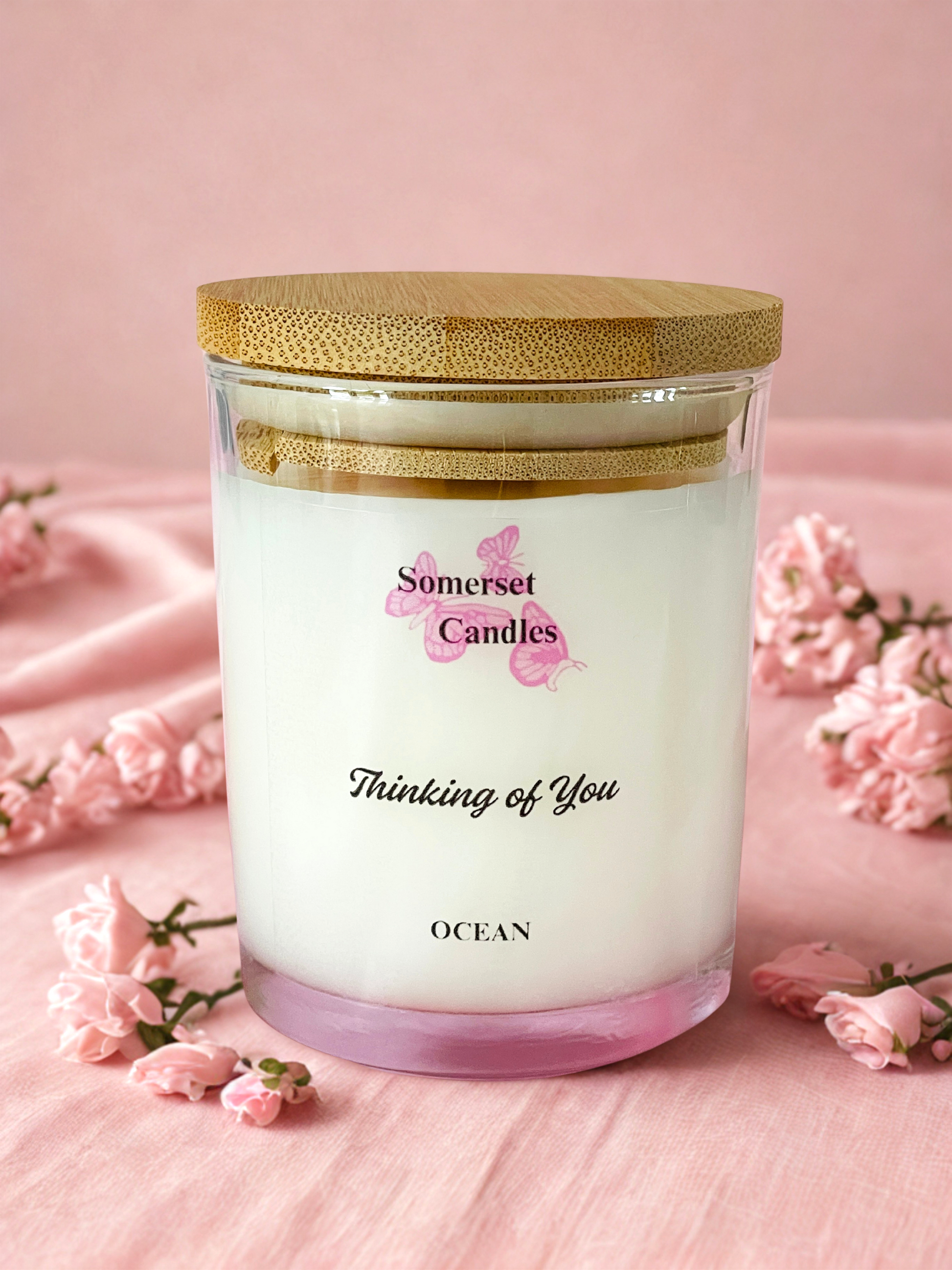 Somerset Candles 'Thinking of You' candle with a wooden lid on a pink fabric background with flowers.