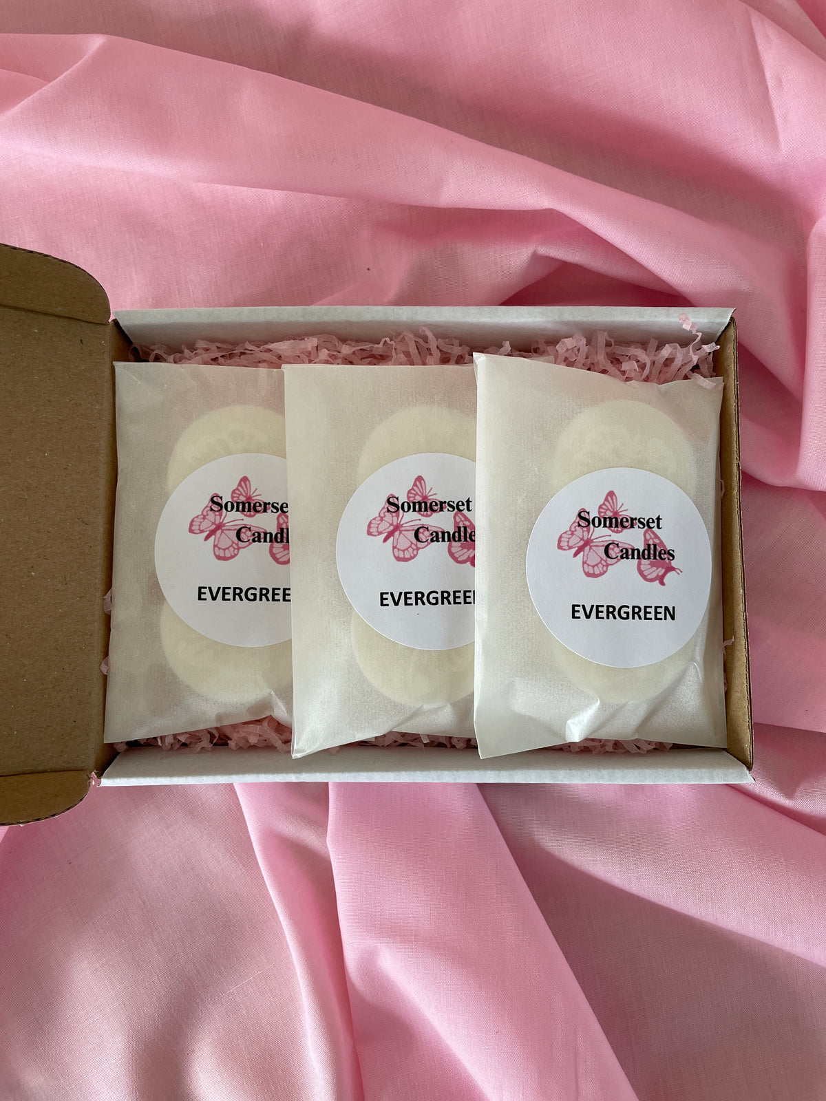 Three individually wrapped wax melts in a white gift box with pink shredded paper, set against a draped pink fabric background. The labels read 'Somerset Candles' and 'EVERGREEN'. Handmade gifts.