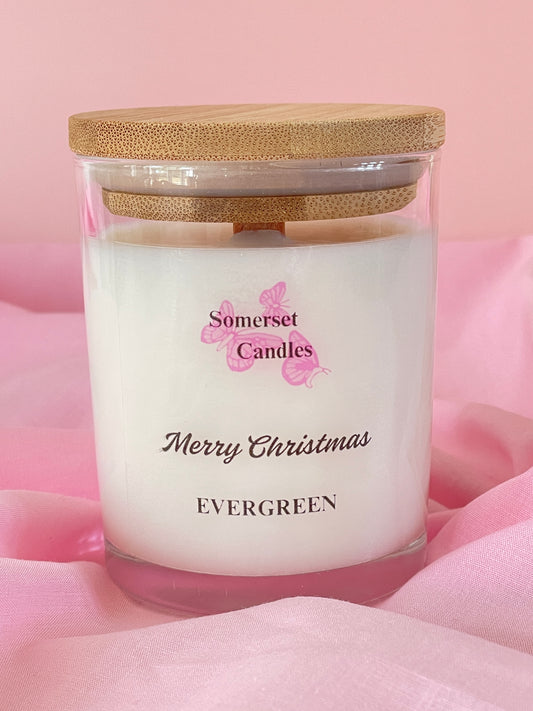 A glass candle jar with a wooden lid. The candle has a white wax interior and a label with the text "Somerset Candles" and "Merry Christmas Evergreen". The jar is set against a pink fabric background.
