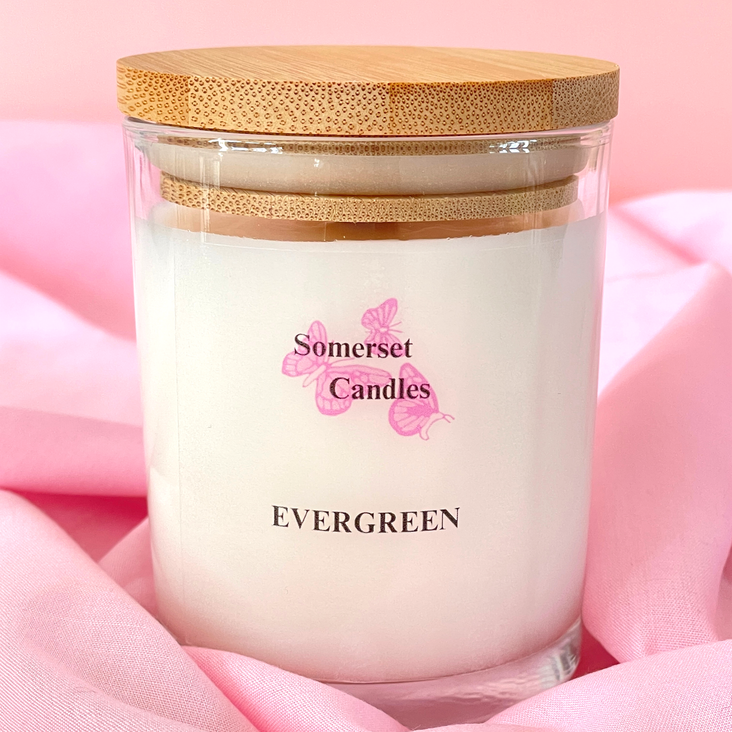 Candle in a glass jar with a wooden lid on a pink fabric background, branded 'Somerset Candles'.