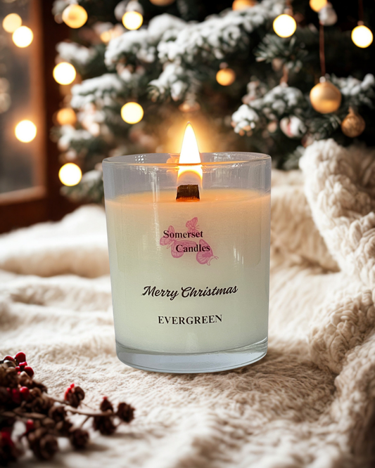 A glass candle jar with a wooden lid. The candle has a white wax interior and a label with the text "Somerset Candles" and "Merry Christmas Evergreen". The jar is set against a cream fabric background and in front of a Christmas Tree. The candle is lit.