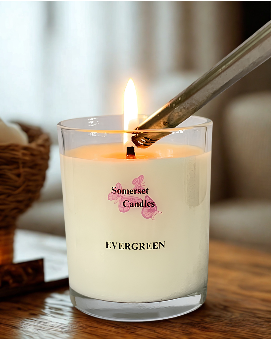 A lit glass jar candle labeled 'Somerset Candles EVERGREEN' sits on a wooden table with a metal lighter resting across the rim. The candle wax is creamy off-white, and the background is softly blurred in a warm indoor setting.