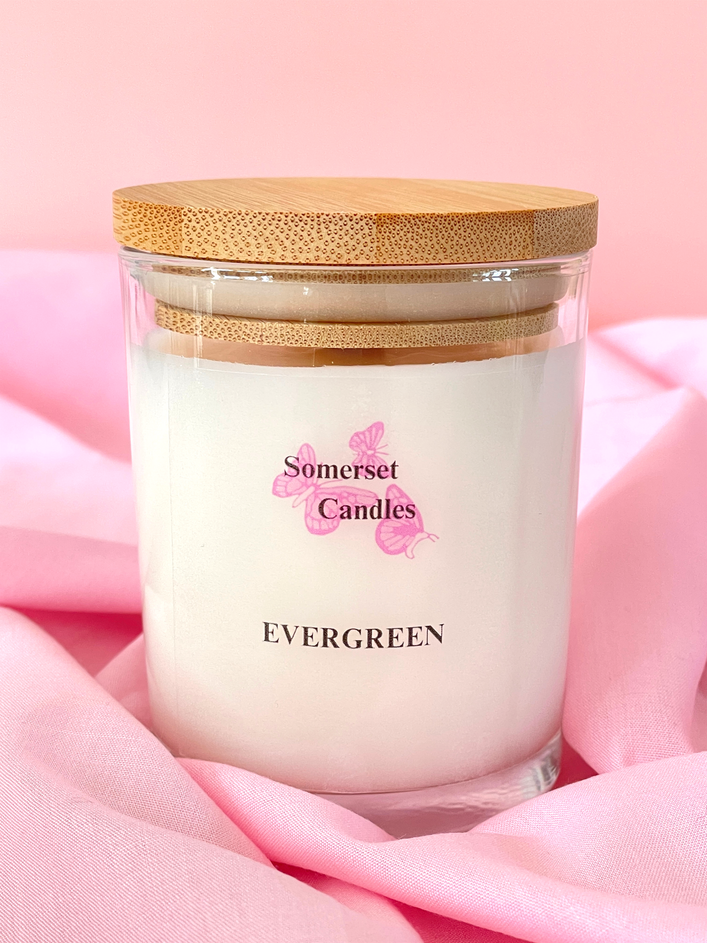 Candle in a glass jar with a wooden lid on a pink fabric background, branded 'Somerset Candles'.