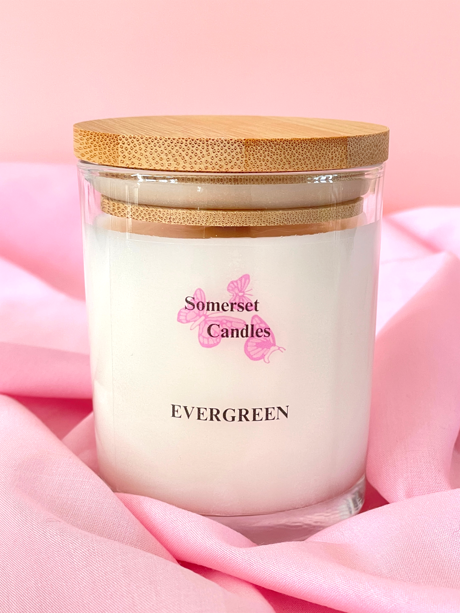 Candle in a glass jar with a wooden lid on a pink fabric background, branded 'Somerset Candles'.