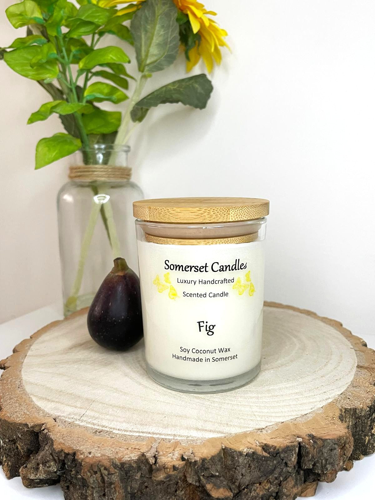 White soy candle in a 7oz clear glass jar with a natural wooden lid. The candle is made from a soy-coconut blend and scented with a fresh scent of ripe fig. Above the scent name on the label, several yellow butterflies are displayed, adding a romantic touch. Clean, minimalist design ideal for gifts, home decor or self-care.