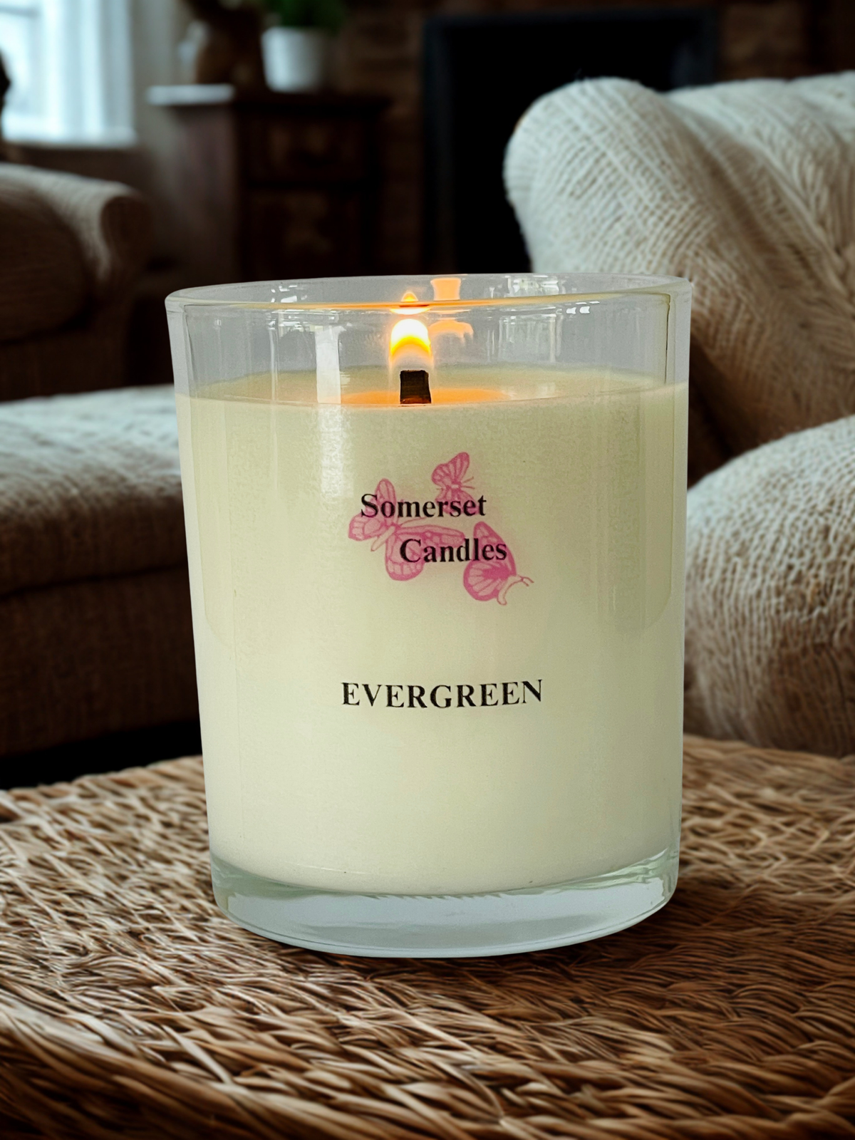 A lit Somerset Candles 'Evergreen' scented candle in a clear glass jar, set on a woven wicker surface with a cozy living room background.