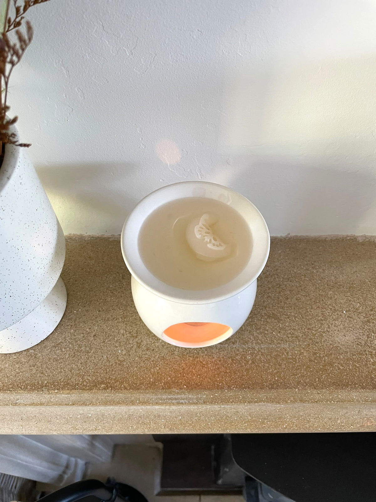 A white ceramic wax warmer candle with a visible orange accent on the front, sitting on a textured beige shelf against a white wall.