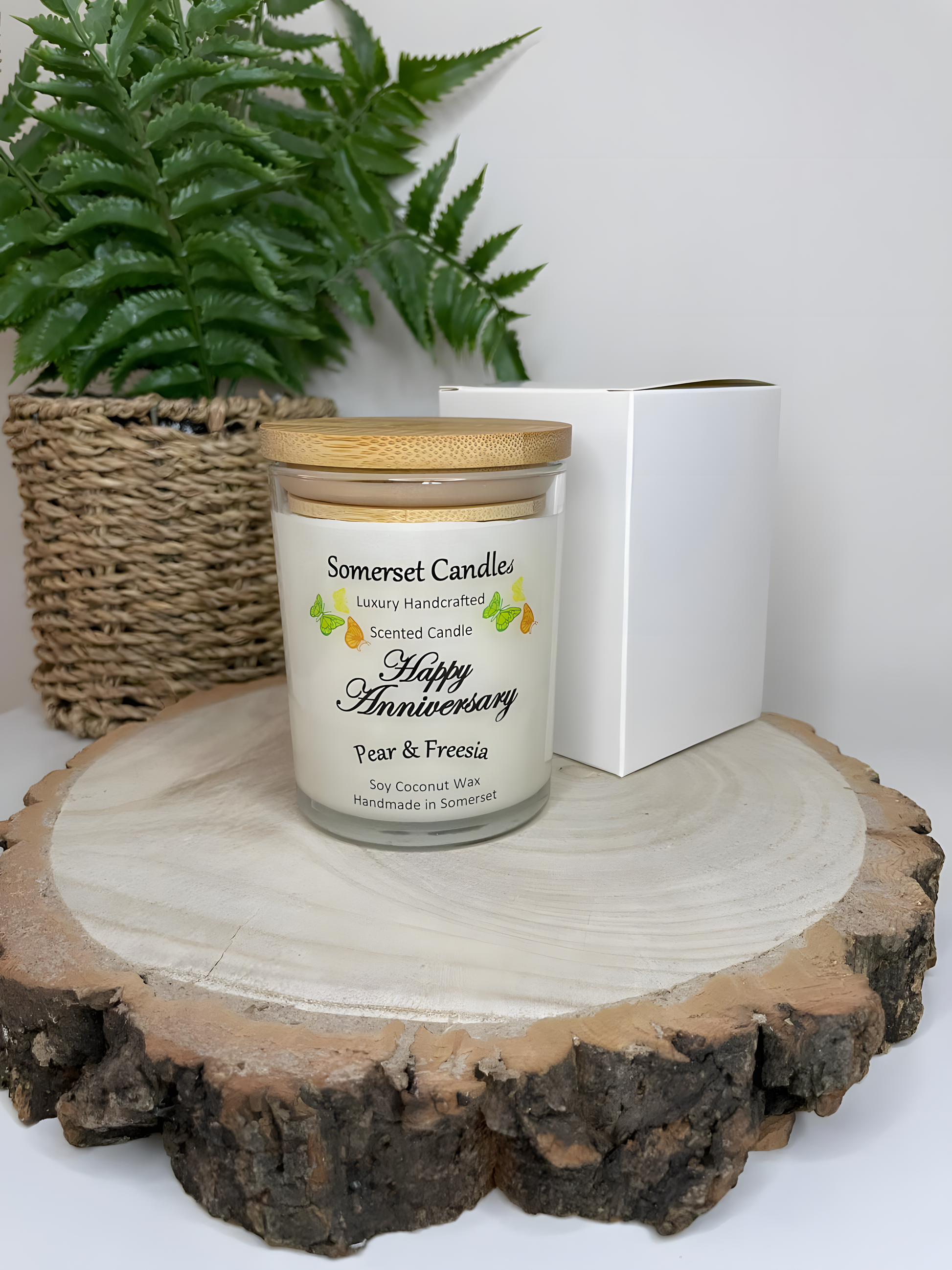 Happy Anniversary Pear & Freesia scented soy candle displayed on a wooden board with a white gift box. The candle is white in colour with bright yellow, green & orange butterflies fluttering above the scent name and fitted with a lovely bamboo lid.