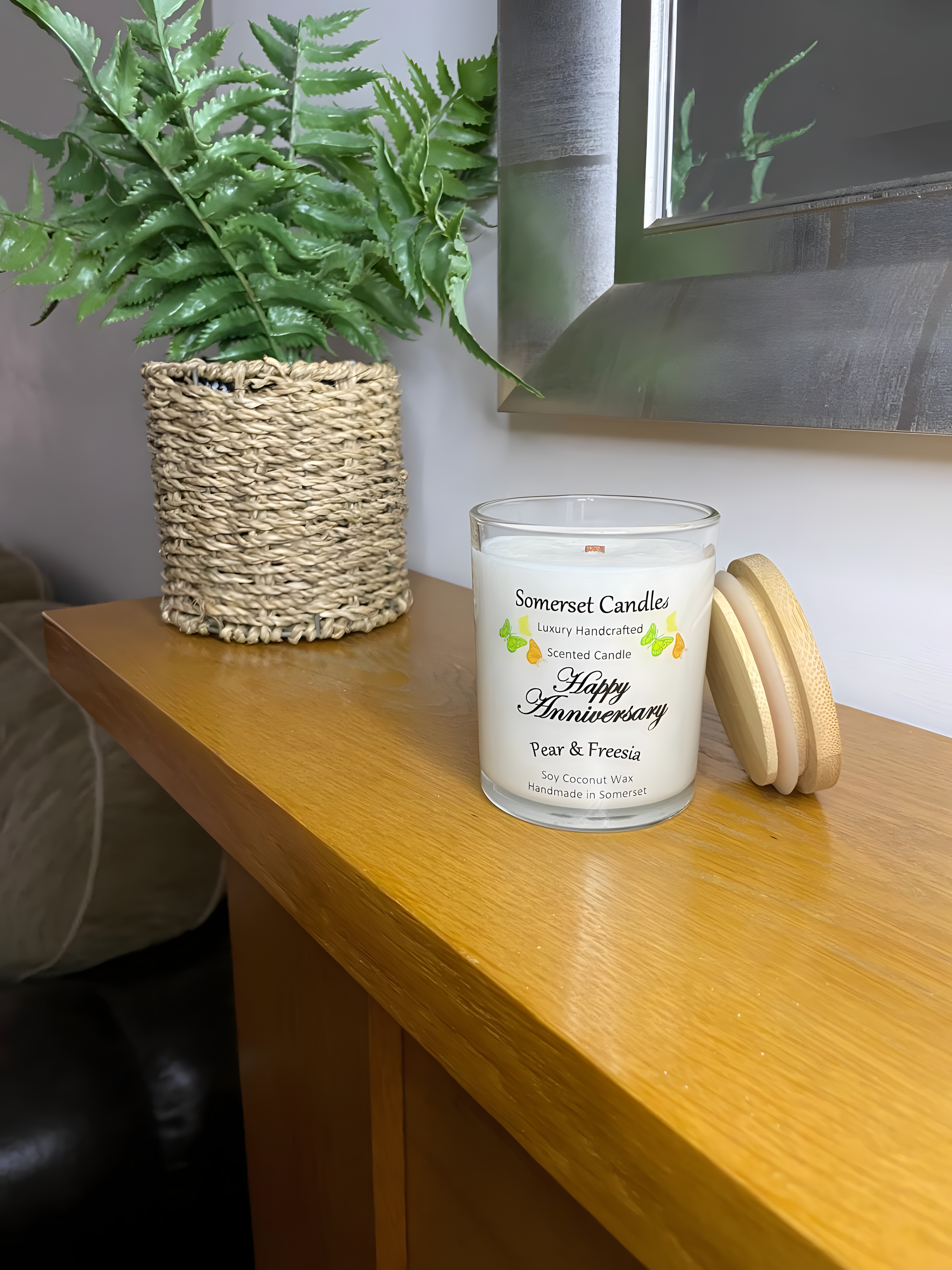 Happy Anniversary Pear & Freesia scented soy candle  on the mantelpiece. The candle is white in colour with bright yellow, green & orange butterflies fluttering above the scent name with a bamboo lid.