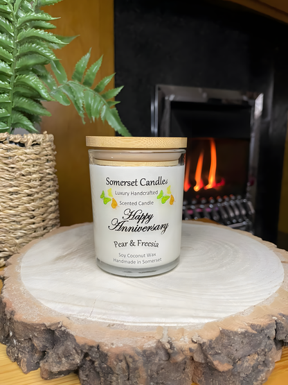 Happy Anniversary Pear & Freesia scented soy candle displayed on a wooden board in front of a lit fire. The candle is white in colour with bright yellow, green & orange butterflies fluttering above the scent name and fitted with a lovely bamboo lid.