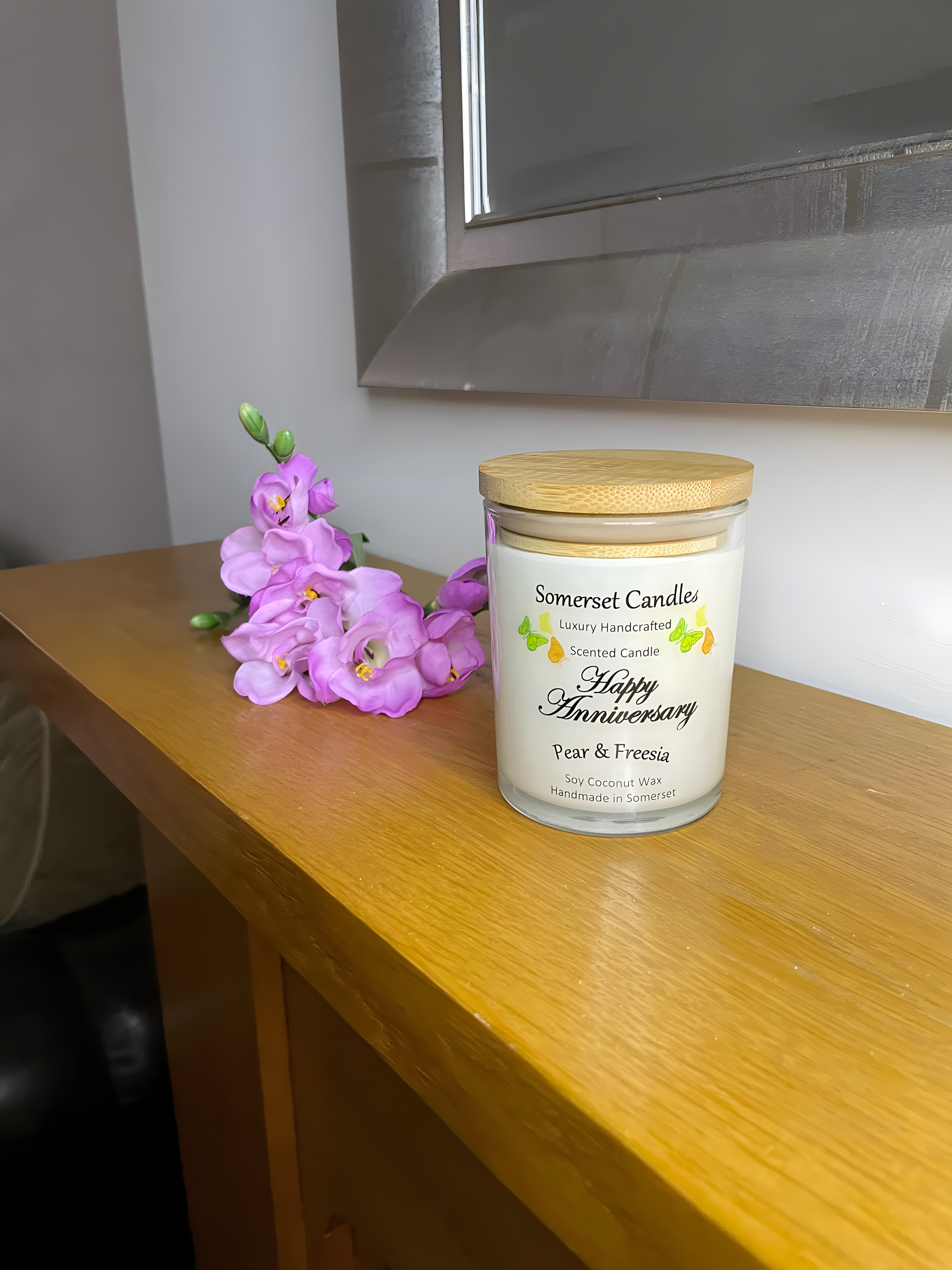 Happy Anniversary Pear & Freesia scented soy candle displayed on the mantelpiece with a freesia. The candle is white in colour with bright yellow, green & orange butterflies fluttering above the scent name and fitted with a lovely bamboo lid.