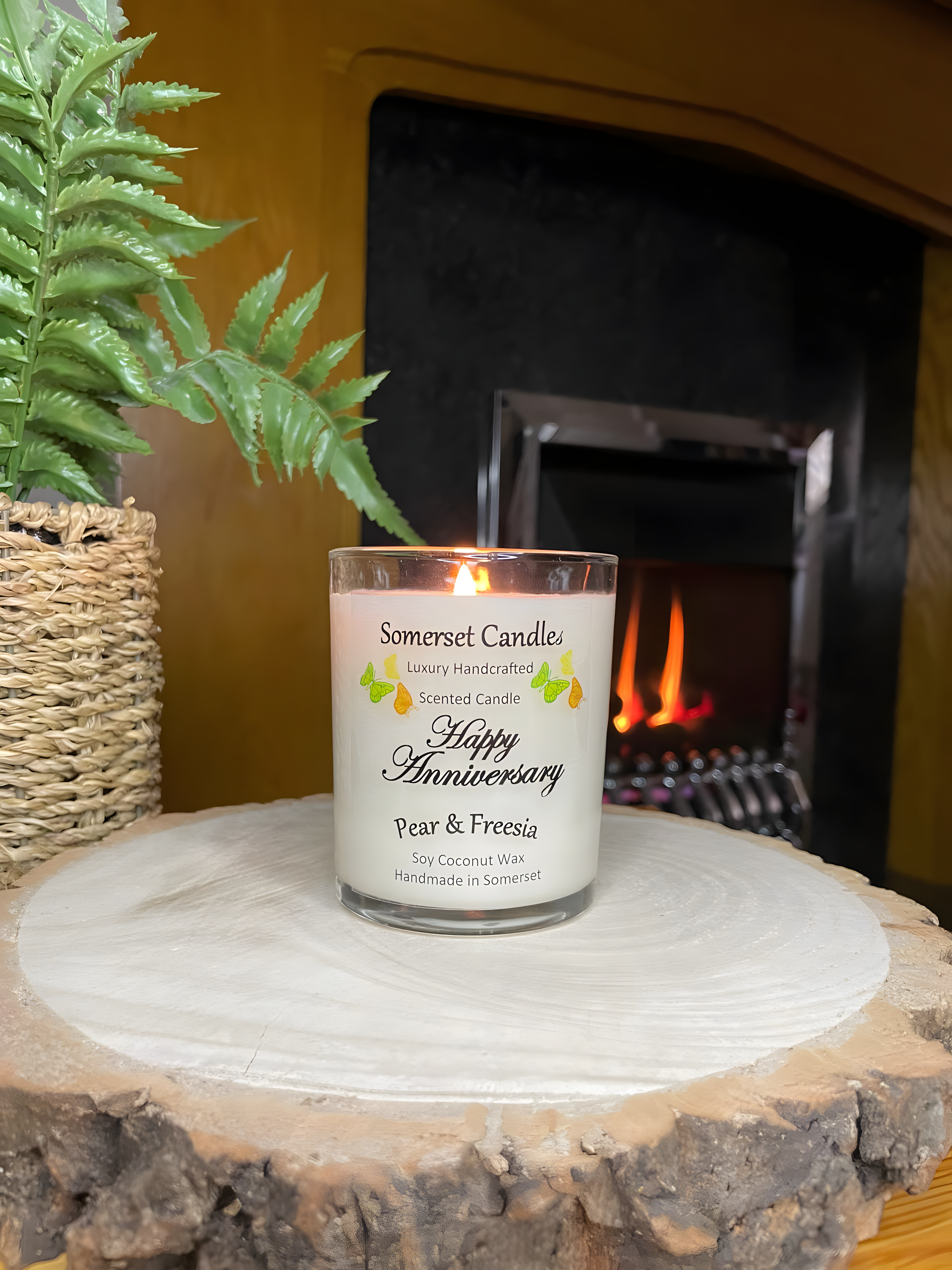 Happy Anniversary Pear & Freesia scented soy candle displayed on a wooden board in front of a lit fire. The candle is white in colour with bright yellow, green & orange butterflies fluttering above the scent name. The candle is lit.