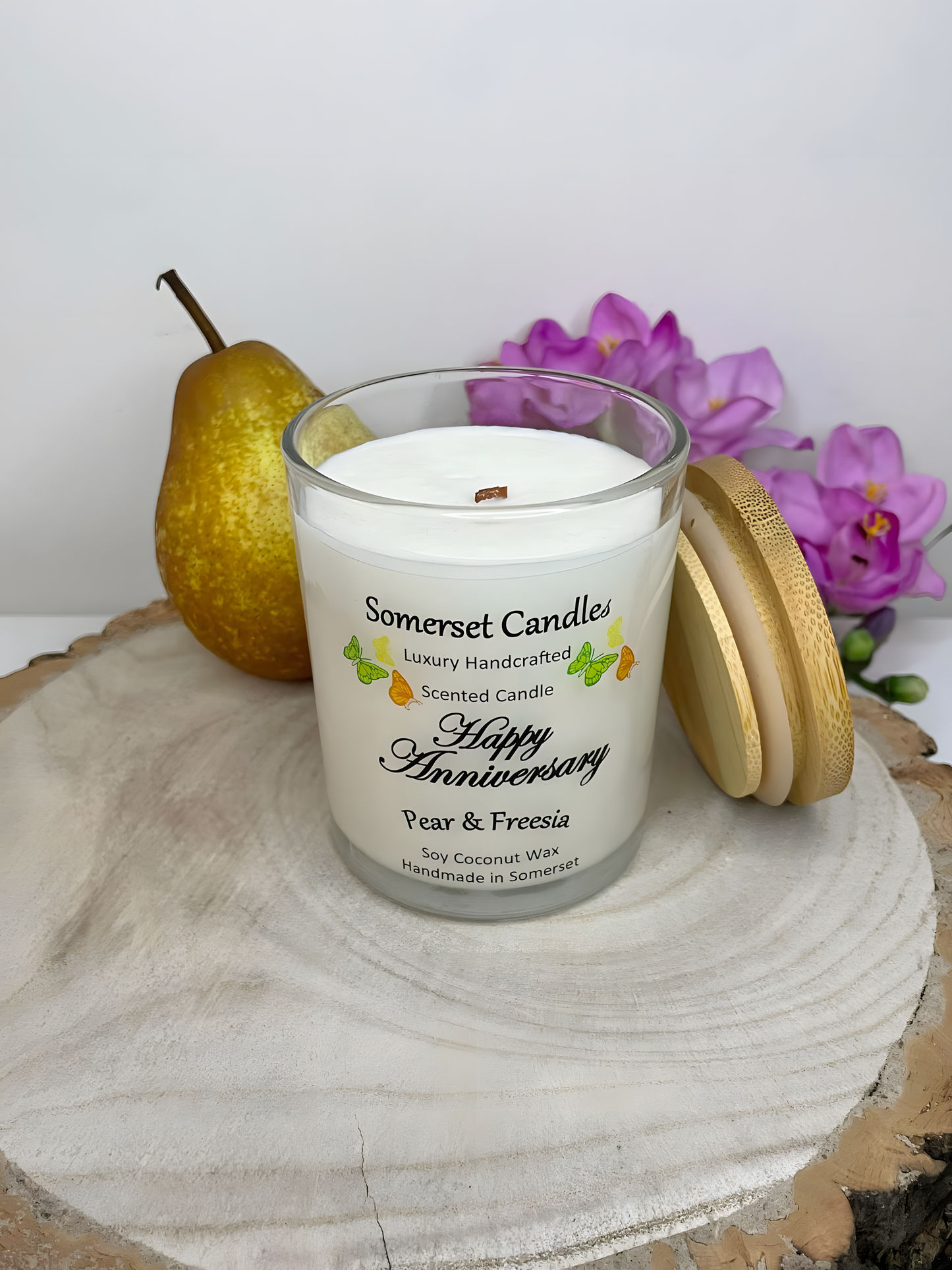 Happy Anniversary Pear & Freesia scented soy candle displayed on a wooden board with fresh pear & freesia. The candle is white in colour with bright yellow, green & orange butterflies fluttering above the scent name and a bamboo lid.