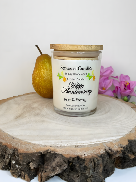 Happy Anniversary Pear & Freesia scented soy candle displayed on a wooden board with fresh pear & freesia. The candle is white in colour with bright yellow, green & orange butterflies fluttering above the scent name and a lovely bamboo lid.