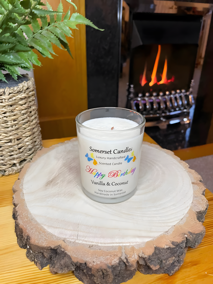 Happy Birthday Vanilla & Coconut scented soy candle displayed on a wooden board in front of a lit fire. The candle is white in colour with bright blue, yellow & orange butterflies fluttering above the scent name. The wooden wick can be seen.