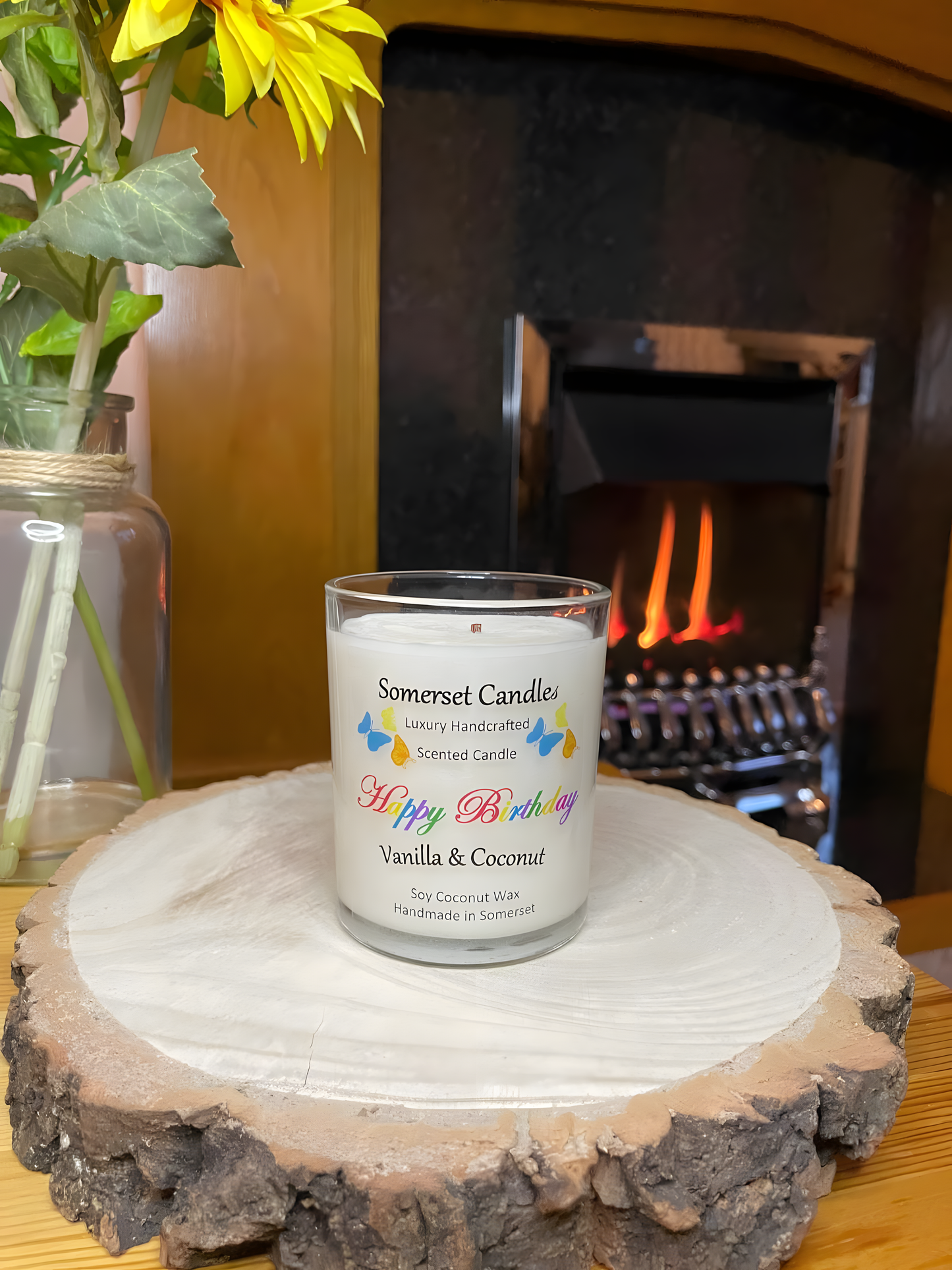 Happy Birthday Vanilla & Coconut 
scented soy candle displayed on a wooden board in front of a lit fire. The candle is white in colour with bright blue, yellow & orange butterflies fluttering above the scent name.