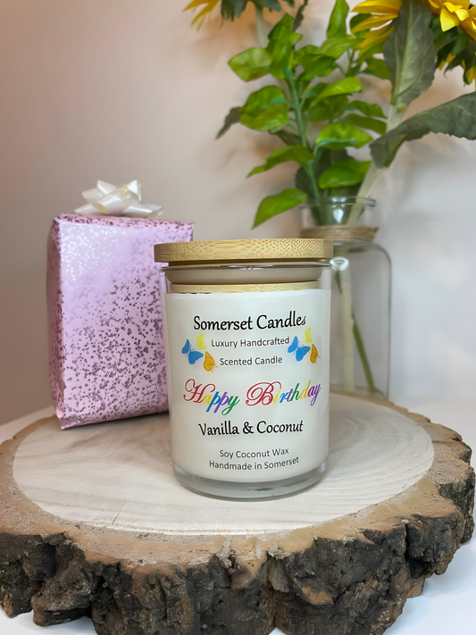 Happy Birthday Vanilla & Coconut scented soy candle displayed on a wooden board with a wrapped gift box. The candle is white in colour with bright blue, yellow & orange butterflies fluttering above the scent name and fitted with a lovely bamboo lid.