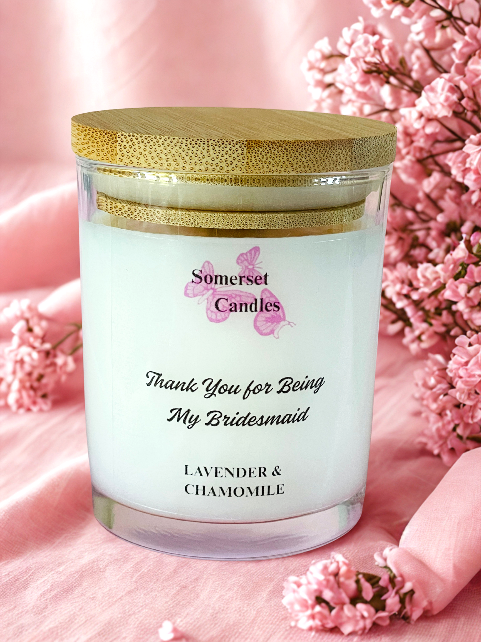 Scented candle with 'Somerset Candles' branding and floral text, surrounded by pink flowers on a soft pink background.
