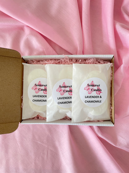 Three individually wrapped wax melts labelled 'Somerset Candles Lavender & Chamomile' arranged in a white gift box with pink shredded filler, set on pink satin fabric.