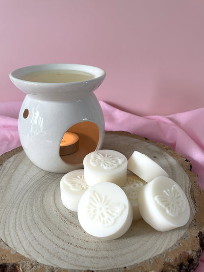 White wax melts with embossed butterfly designs arranged on a rustic wood slice next to a white ceramic candle warmer with a tea light, set against a soft pink fabric backdrop.