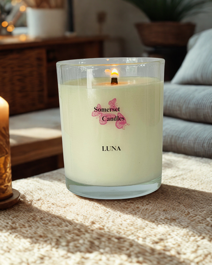 A lit 'Somerset Candles' jar candle named 'LUNA' with pale cream wax, set on a textured beige blanket in a cozy, softly blurred living room environment.