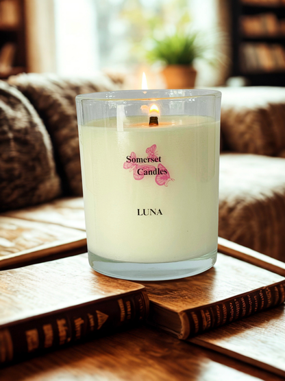A lit candle in a clear glass jar sits on a wooden table in a cozy indoor setting. The candle wax is creamy off-white, with the brand name 'Somerset Candles' and scent name 'LUNA' printed on the front.
