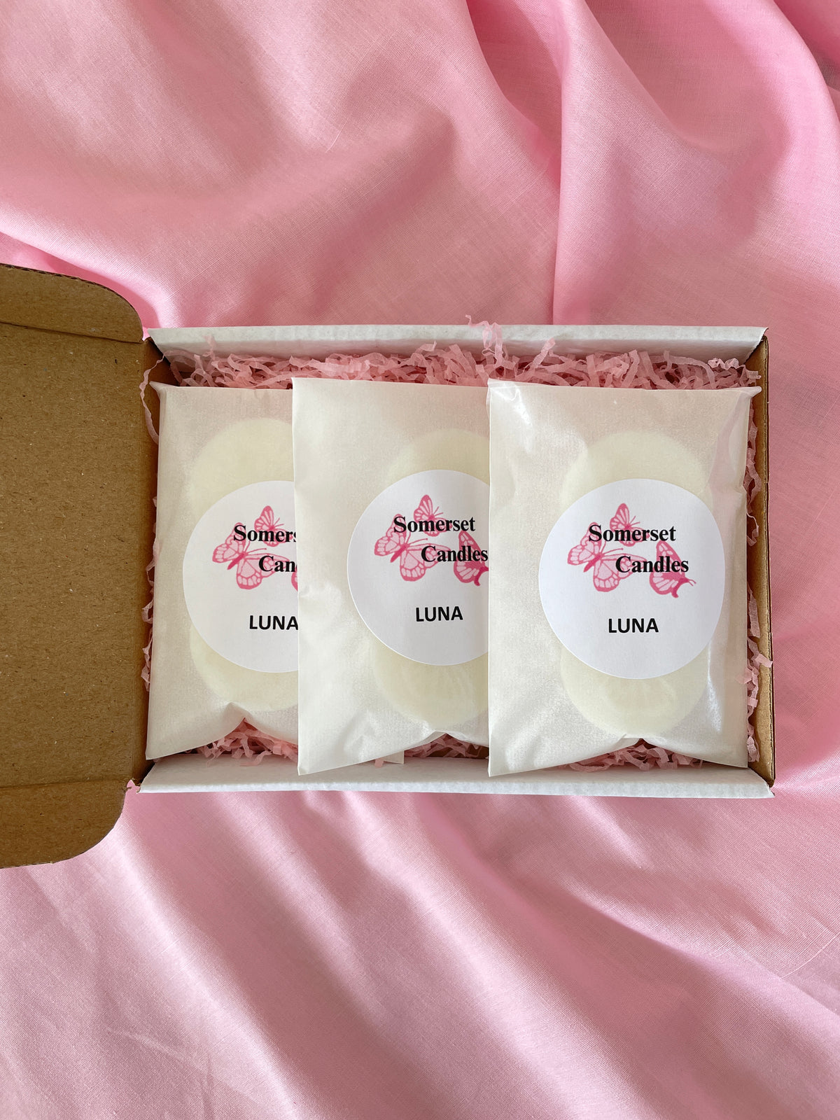Three round wax melts in clear sleeves with white labels reading 'Somerset Candles LUNA' are arranged in a white gift box filled with pink shredded paper, set on pink satin fabric.