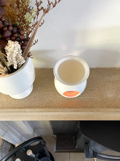 A white ceramic wax warmer with a small orange accent on the front, sitting on a beige stone shelf next to a white vase with dried flowers.