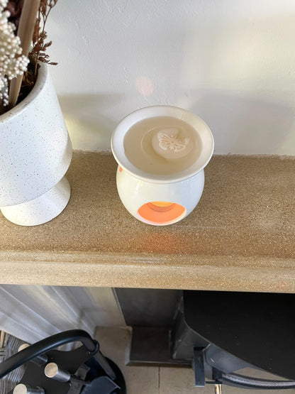 A white ceramic wax warmer candle with a glowing orange base sits on a beige stone shelf next to a white speckled vase with dried flowers.