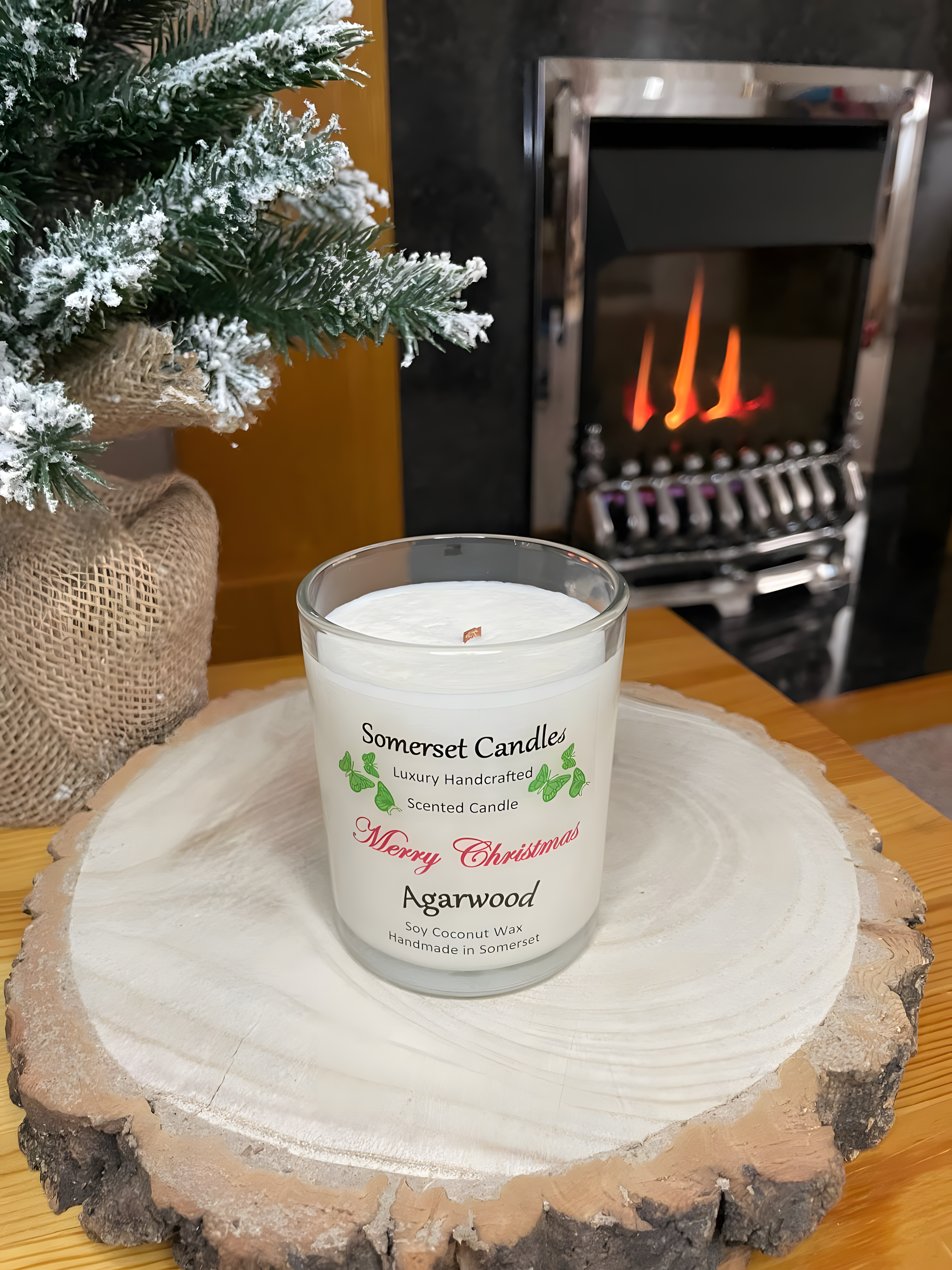 Merry Christmas Agarwood soy candle is displayed on a wooden board in front of a lit fire. The candle is white in colour with bright green butterflies above the scent name and has no lid fitted. The tip of the wick can be seen.