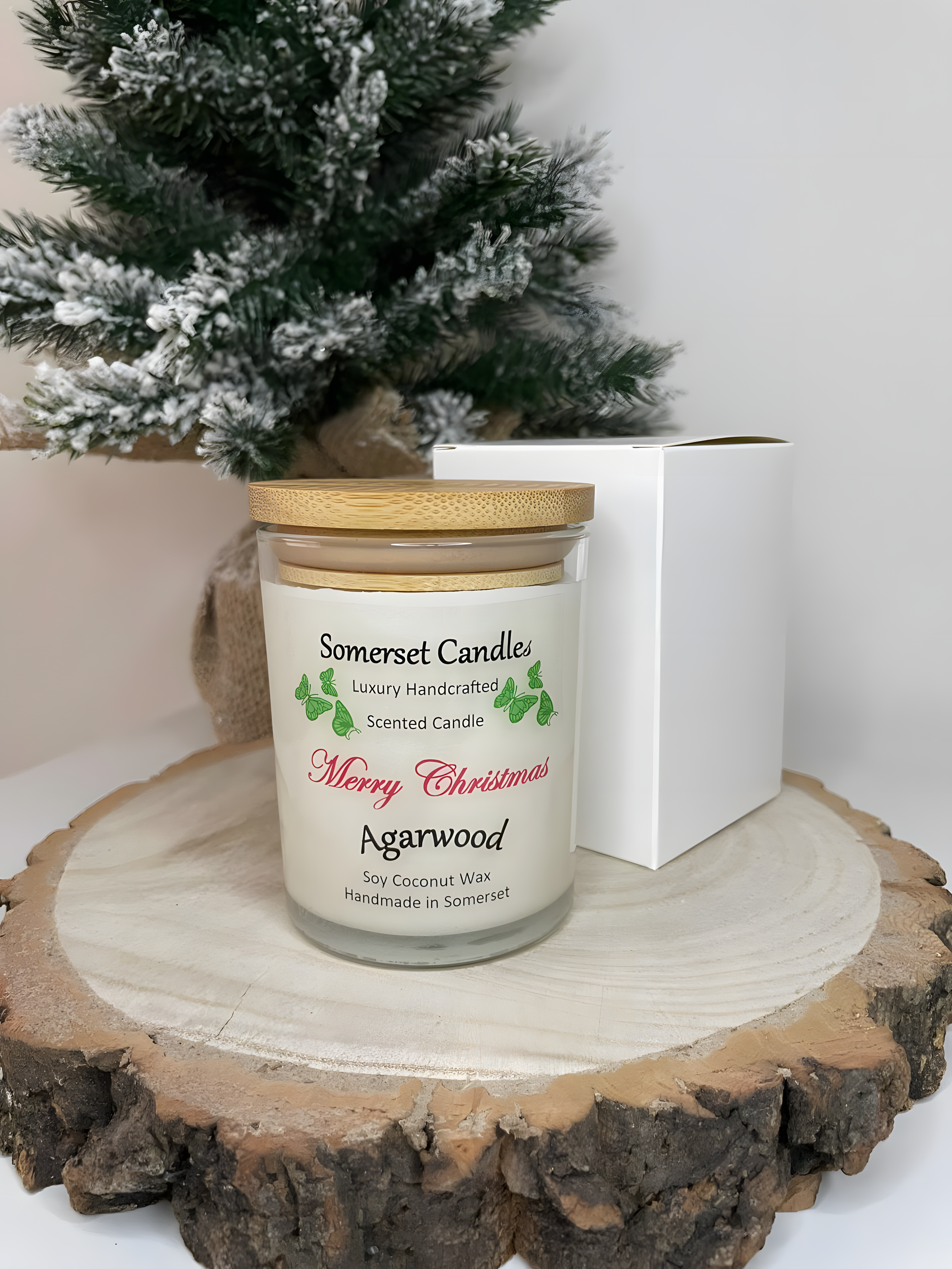 Merry Christmas Agarwood soy candle is displayed on a wooden board with a white gift box and a plant behind. The candle is white in colour with bright green butterflies above the scent name and a bamboo lid.