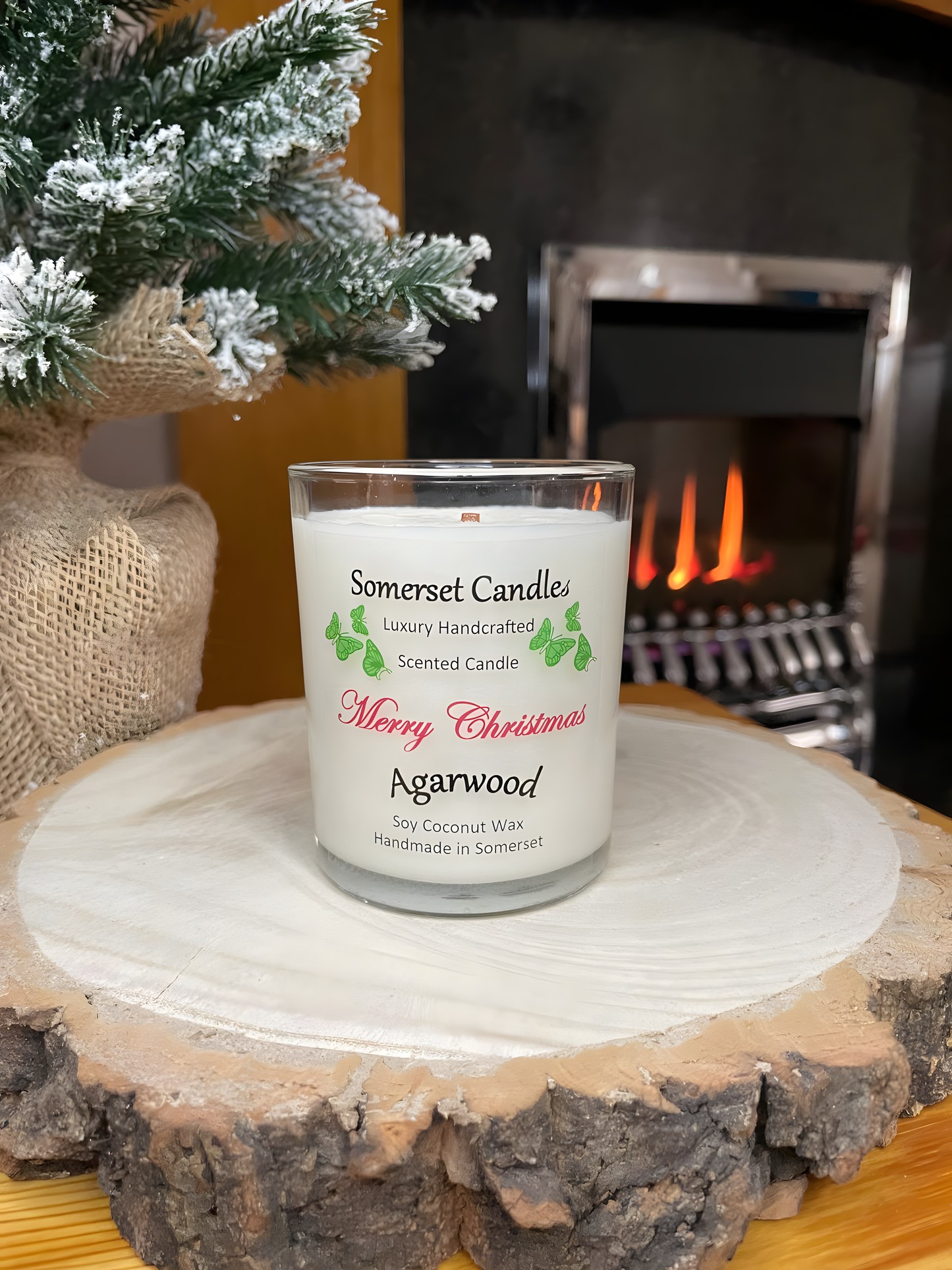 Merry Christmas agarwood soy candle is displayed on a wooden board with a plant behind in front of a lit fire. The candle is white in colour with bright green butterflies above the scent name and with no lid fitted. The tip of the wick can be seen.