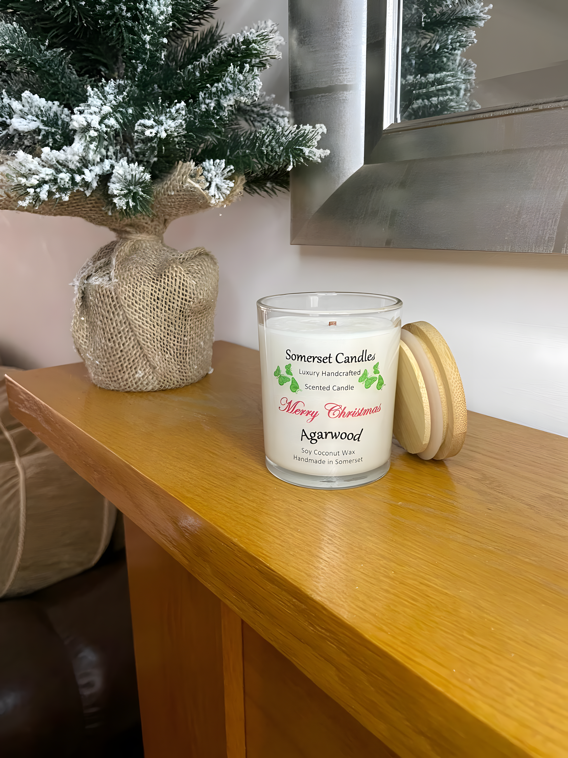 Merry Christmas Agarwood soy candle is displayed on a wooden mantel piece of a fire with a small Christmas tree behind. The candle is white in colour with bright green butterflies above the scent name and a bamboo lid resting against the candle.