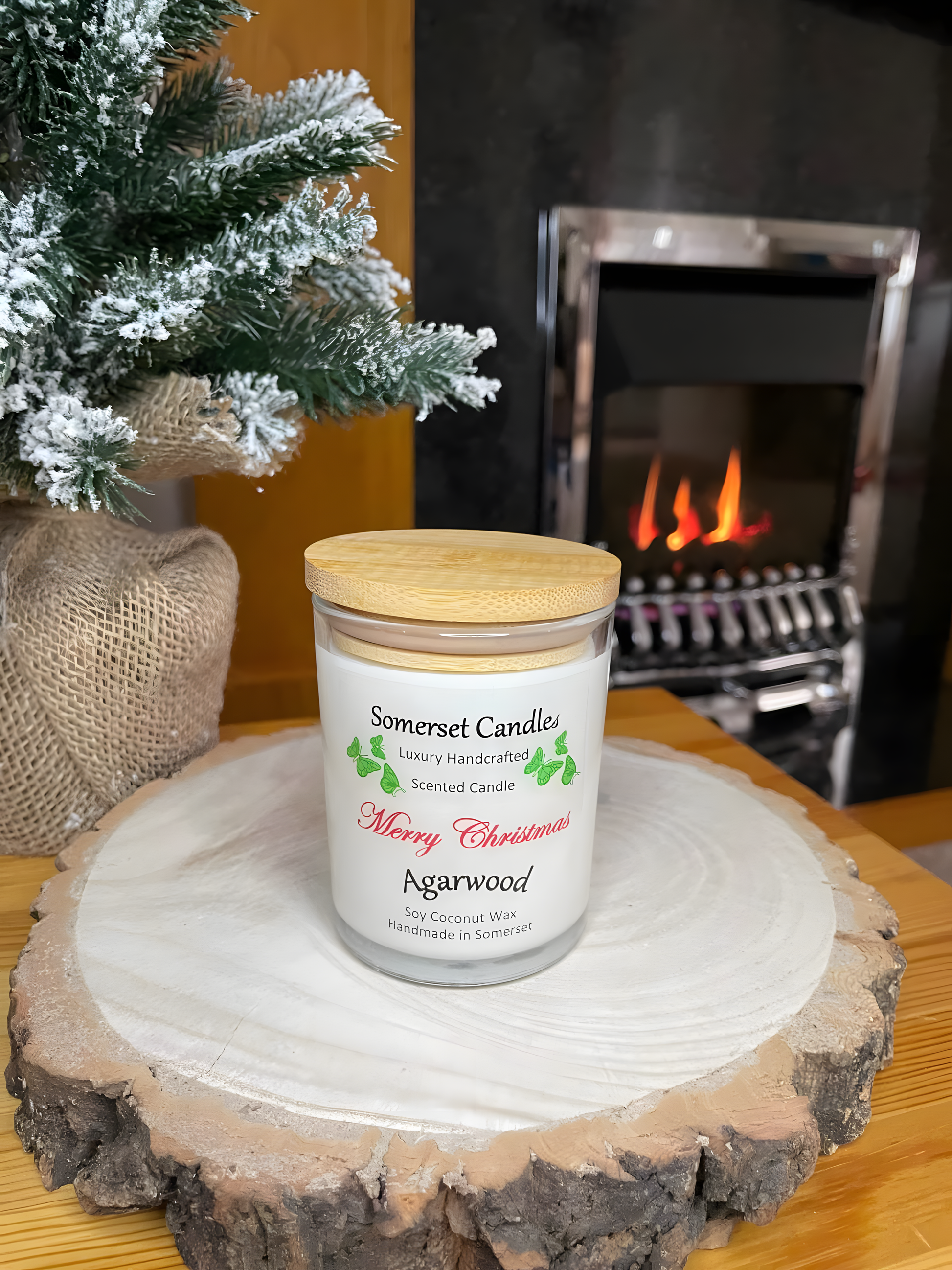 Merry Christmas Agarwood soy candle is displayed on a wooden board with a small Christmas tree behind, in front of a lit fire. The candle is white in colour with bright green butterflies above the scent name and a bamboo lid.