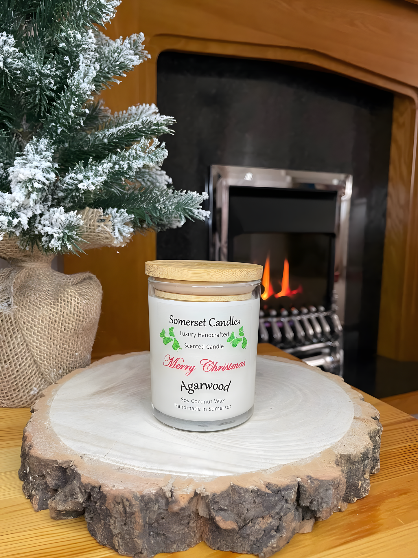 Merry Christmas Agarwood soy candle is displayed on a wooden board with a small Christmas tree behind, in front of a lit fire. The candle is white in colour with bright green butterflies above the scent name and a bamboo lid.