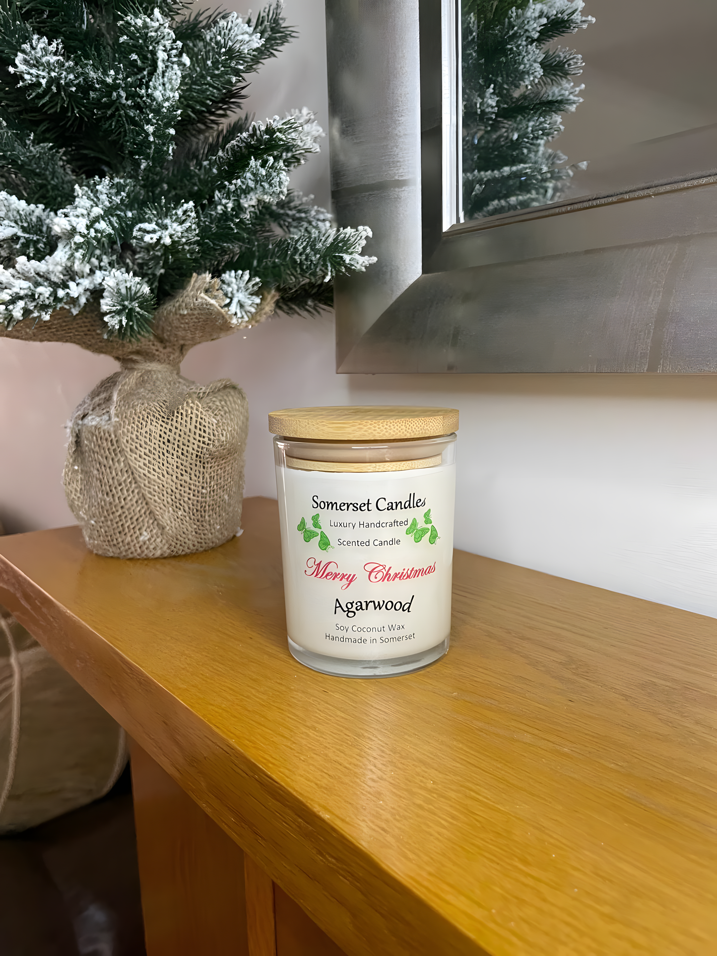 Merry Christmas Agarwood soy candle is displayed on a wooden mantel piece of a fire with a small Christmas tree behind. The candle is white in colour with bright green butterflies above the scent name and a bamboo lid.