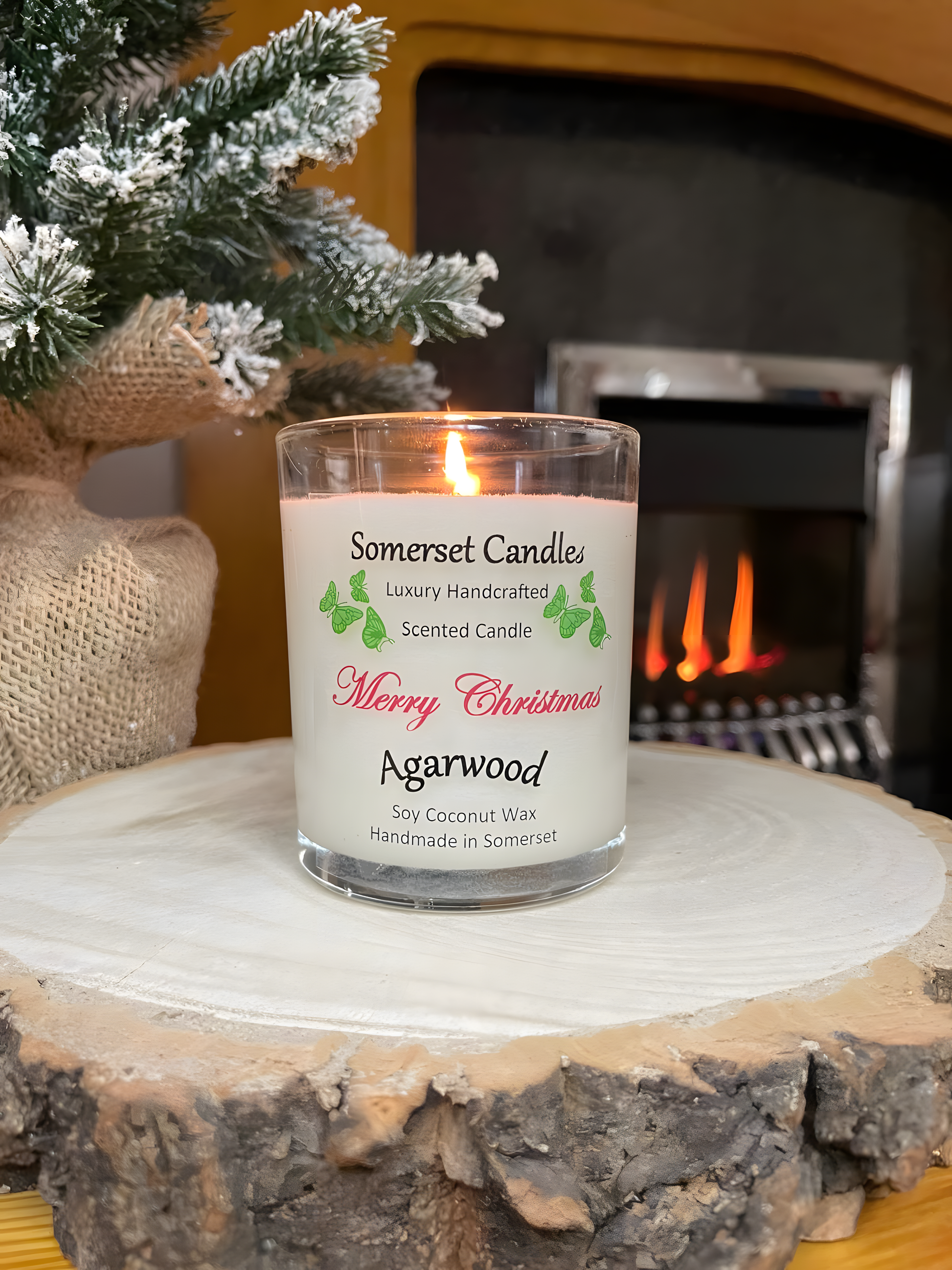 Merry Christmas Agarwood soy candle is displayed on a wooden board in front of a lit fire with a small Christmas tree to the left behind it. The candle is white in colour with bright green butterflies above the scent name and the candle is lit.