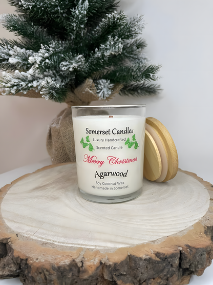 Merry Christmas Agarwood soy candle is displayed on a wooden board with a small Christmas tree behind. The candle is white in colour with bright green butterflies above the scent name and a bamboo lid resting against the candle.