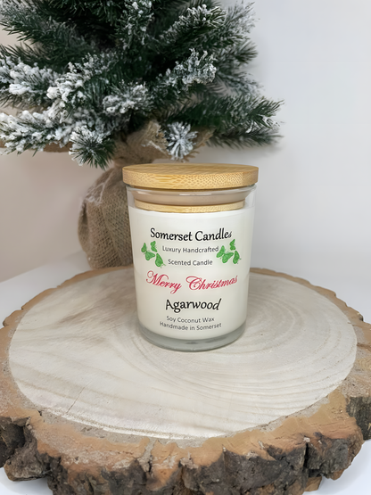 Merry Christmas agarwood soy candle is displayed on a wooden board with a plant behind. The candle is white in colour with bright green butterflies above the scent name and a bamboo lid.