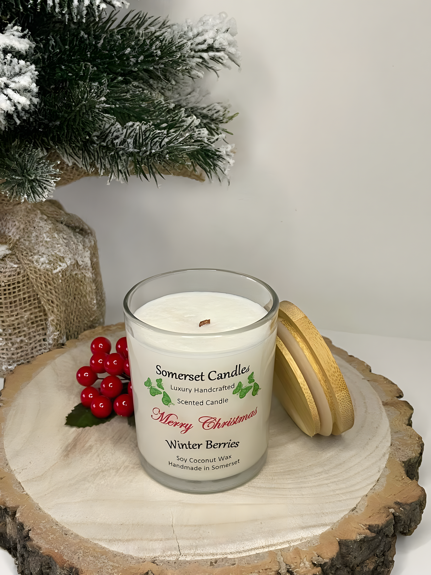 Christmas Winter Berries soy candle is displayed on a wooden board with berries and a small Christmas tree to the left behind. The candle is white in colour with bright green butterflies above the scent name and has a bamboo lid leaning on the candle